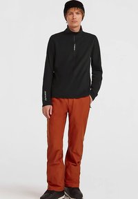 Sort fleece-top med lynlås og logo på ærmet, kombineret med orange vandtætte bukser, båret med sorte sko og en sort hue.