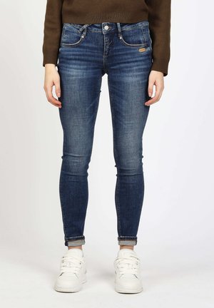 Person trägt dunkelblaue Skinny-Jeans, braunen Pullover und weiße Turnschuhe, steht mit entspannten Händen an den Seiten.