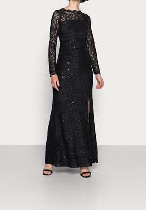 Kobieta w długiej czarnej koronkowej sukni wieczorowej z długimi przezroczystymi rękawami, rozcięciem z przodu i czarnymi, otwartymi butami na obcasie.