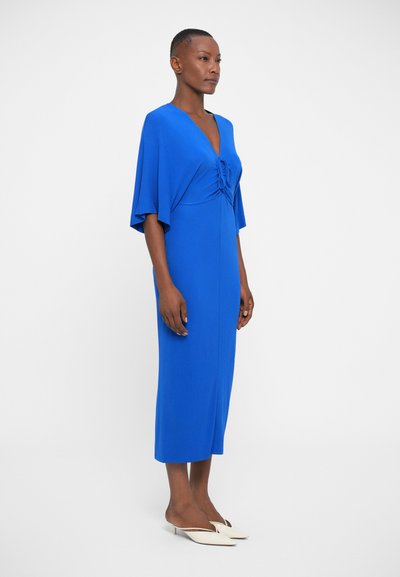 Modré midi šaty s výstřihem ve tvaru V a draped stylovými rukávy. Hladká látka s nařaseným detailem na hrudi. Kombinováno s bílými mules na podpatku.