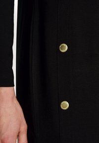 Černý oděv s hladkou texturou, dlouhými rukávy a zlatými knoflíkovými detaily podél bočního švu. Ruka viditelná, jak drží látku.
