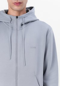 Sudadera con cremallera de color gris claro, hecha de una tela suave y elástica. Cuenta con capucha con cordón, bolsillos laterales y logo "BOSS" en relieve.