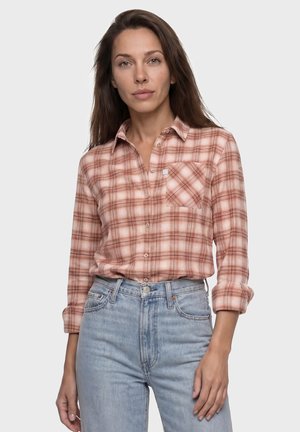 Frau mit langen braunen Haaren, die ein pink kariertes Hemd mit hochgekrempelten Ärmeln und hellblaue, hoch taillierte Jeans vor einem schlichten Hintergrund trägt.