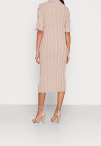 Robe midi en tricot côtelé beige avec des manches courtes, présentant un col replié et un ourlet festonné. Portée avec des sandales à talons nus.