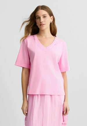 Pink cotton t-shirt with a V-neck and short sleeves, featuring subtle text embroidered around the neckline, layered over a pink skirt.