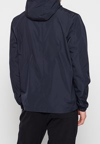 Homme portant une veste imperméable à capuche bleu foncé vue de dos, associée à un pantalon noir.