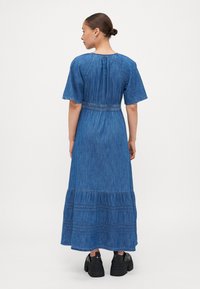Denim maxikjole med korte ermer, samlet midje og volangkant. Laget av lett blått stoff, med horisontale sømdetaljer.