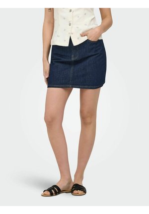 Femme portant une mini-jupe en jean bleu foncé, un haut sans manches léger à motifs et des sandales plates noires, debout avec une main dans la poche de la jupe.