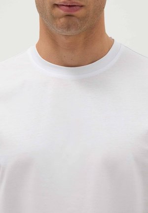 Nahaufnahme des unteren Gesichts und Halses eines Mannes, der ein einfaches weißes T-Shirt mit Rundhalsausschnitt trägt, vor neutralem Hintergrund.