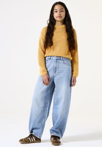 Pull jaune tricoté léger avec une maille lâche et ouverte. Associé à un jean baggy bleu clair taille haute et des baskets à motifs marron.