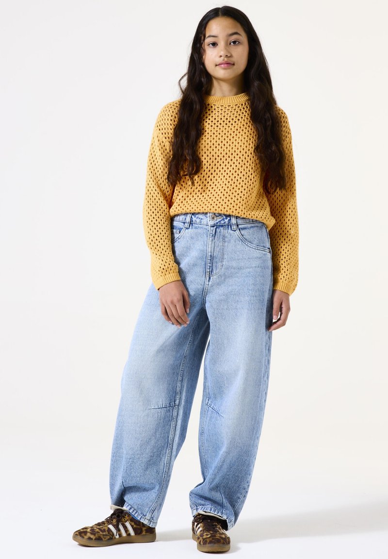 Pull jaune tricoté léger avec une maille lâche et ouverte. Associé à un jean baggy bleu clair taille haute et des baskets à motifs marron.