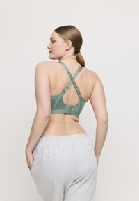 Mujer con cabello castaño claro en una coleta, vistiendo un sujetador de tirantes cruzados de color verde claro y pantalones de chándal grises claros, con las manos en los bolsillos.