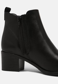Bottine noire en cuir avec fermeture éclair latérale, talon bloc et languette à l'arrière, présentée sur un fond clair uni.