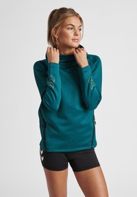 Türkisfarbener Pullover mit langen Ärmeln, hohem Kragen, Reißverschlusstaschen an den Seiten und kontrastierenden goldenen Akzenten an den Ärmeln. Glattes, elastisches Material.