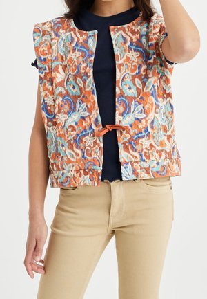 Donna che indossa un gilet senza maniche arancione con motivo floreale sopra una maglia nera e pantaloni beige, con un braccio sollevato vicino alla testa.