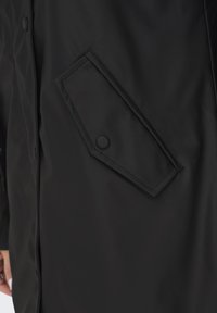 Manteau noir avec une poche à rabat inclinée sécurisée par un bouton-pression, et une partie de la main visible sur le côté.