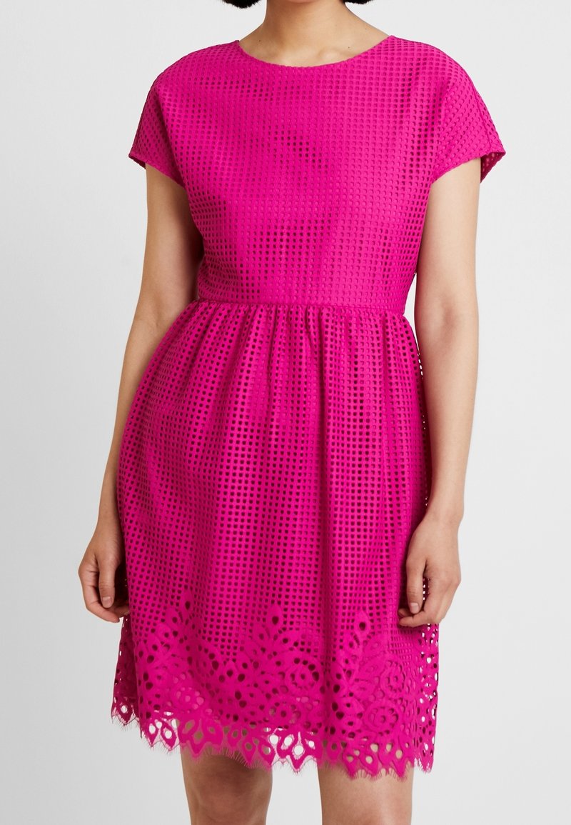 Femme portant une robe sans manches rose vif avec un motif quadrillé texturé et des détails de dentelle florale à l'ourlet.