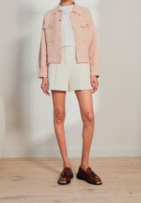 Personne portant une veste à carreaux rose, un haut blanc, un short taille haute crème et des mocassins à franges marron, se tenant sur un sol en bois.