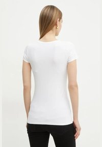 T-shirt en coton blanc à manches courtes, design ajusté, col rond, texture lisse, porté avec un pantalon noir, cheveux coiffés en arrière.