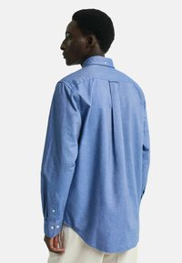 Blå chambray-skjorte med lange ermer, en knapp-ned krage, pleat-detalj bak og avrundet hem. Har knappelukking ved mansjettene.