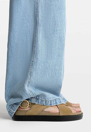 Light blue denim jeans covering feet wearing tan suede sandals with black soles and adjustable buckles on a white surface.
