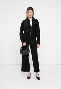Czarna bomberka z cekinami, z zamkiem z przodu, dopasowanymi mankietami, noszona na białej bluzce, w połączeniu z czarnymi spodniami wide-leg i czarną torebką.