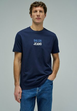 Hombre con pelo rizado que lleva una camiseta azul marino "Salsa Jeans" y vaqueros azules, de pie con una mano en el bolsillo frente a un fondo liso.