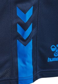Tummansininen kangas, jossa on kolme alas suuntautuvaa sinistä chevron-raitaa ja osittainen sininen "hummel"-urheiluvaatetuslogo oikealla puolella.