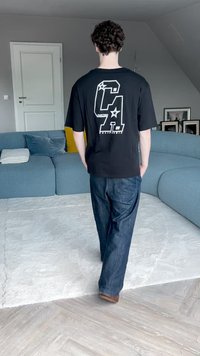 Čierne oversized tričko s bielou grafikou, ktorá zobrazuje písmená a hviezdy na chrbte. Skombinované s voľnými džínsami a hnedými topánkami.
