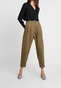 Blusa nera a maniche lunghe con scollo a V, abbinata a pantaloni oliva a vita alta con pieghe e tacchi a motivi bianco e nero.