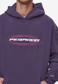 Fialová mikina s kapucí z bavlny s oválným potiskem vpředu obsahujícím nápis "PEGADOR MOST ALIVE" v bílé a růžové barvě. Má kapuci se šňůrkou.