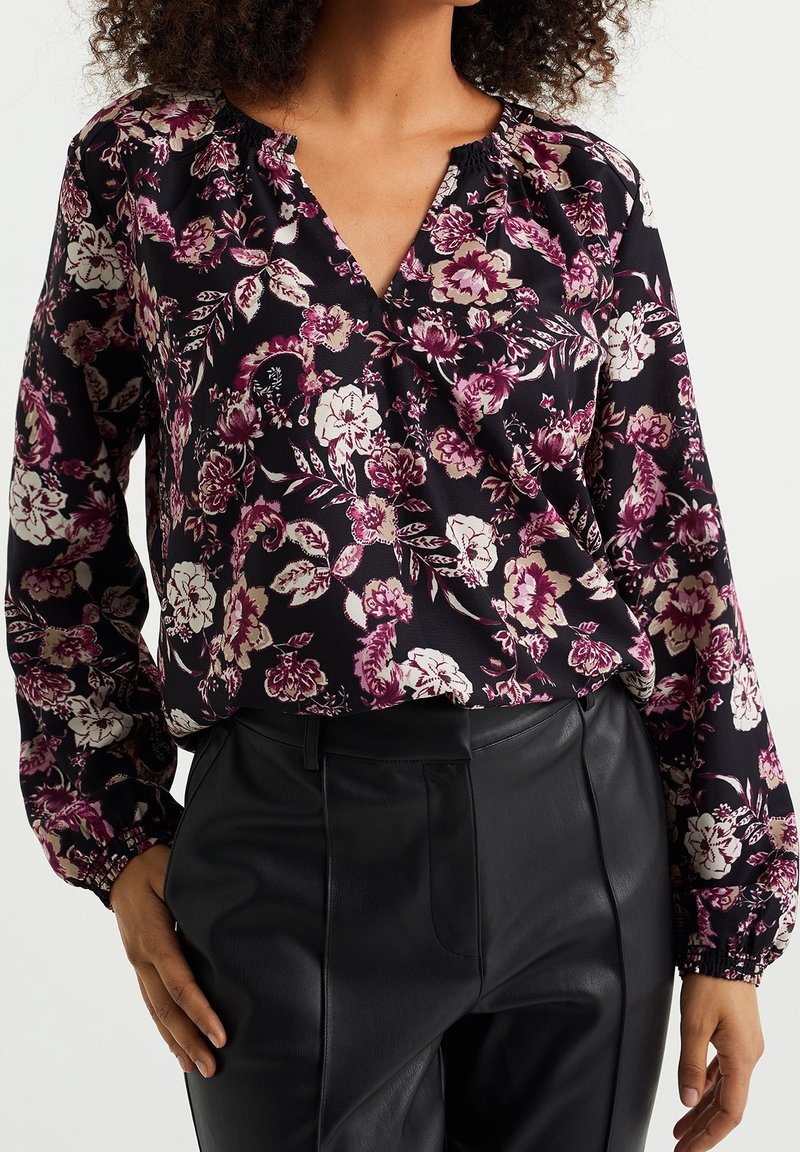 Femme portant un chemisier noir à imprimé floral violet et blanc et un pantalon noir taille haute en cuir synthétique, mains détendues le long du corps.