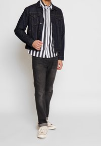 Donkerdenimjack met twee borstzakken, gedragen over een wit en zwart gestreept overhemd met knopen. Vervagende donkere jeans en lichte sneakers maken de outfit compleet.