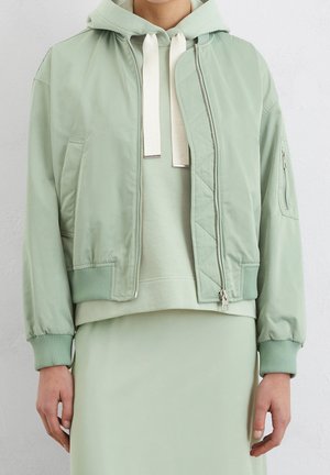 Osoba ubrana w jasnozieloną kurtkę typu bomber zapinaną na zamek, nałożoną na pasującą bluzę z kapturem z białymi ściągaczami i jasnozieloną spódnicę.