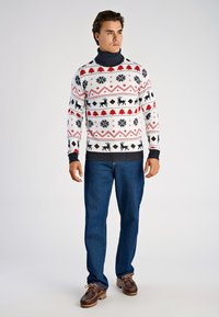 Pull en tricot à col montant, présentant des motifs blancs, rouges et noirs de rennes, flocons de neige et arbres. Associé à un jean bleu et des chaussures marron.