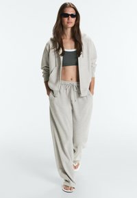 Femme portant une veste zippée à capuche gris clair, un pantalon ample assorti, un soutien-gorge de sport foncé, des lunettes de soleil noires et des tongs blanches, marchant vers l'avant.