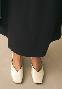 Weiße spitze Schuhe mit Krokodil-Leder-Textur, schwarzem Rand und offenen Seiten, kombiniert mit einem langen schwarzen Kleidungsstück, auf einer hellen Fläche.