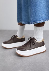 Ruske platform sneakers med teksturert design, hvit såle, gull detaljer og brune lisser, kombinert med hvite ribbestrikkede sokker og et denimskjørt.