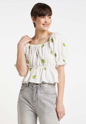 Giovane donna con capelli corti e castani sorride indossando una camicetta bianca con maniche a sbuffo con stampe floreali verdi e jeans a vita alta grigio chiaro.