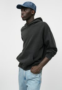 Haut à capuche gris foncé avec une poche avant et une coupe décontractée, associé à un jean bleu clair. La personne porte une casquette à motif bleu.