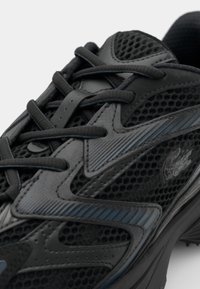 Gros plan d'une chaussure de sport noire avec mesh, surpiqûres synthétiques, lacets épais et petit logo en forme de crocodile sur le côté.