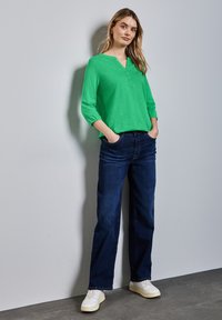 Grön, långärmad henley-tröja med knapplist, kombinerad med mörkblå jeans och vita sneakers. Enkel, avslappnad passform.