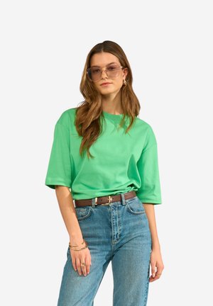 Kvinde iført oversize grøn t-shirt, højtaljede blå jeans, brun bælte, solbriller og guldsmykker, stående mod en hvid baggrund.