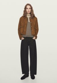 Donna con lunghi capelli ondulati indossa una giacca di suede marrone, un dolcevita a righe, pantaloni larghi neri e scarpe nere, in piedi con le mani nelle tasche.