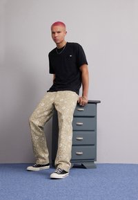 T-shirt nera a maniche corte con taschino, pantaloni beige a fantasia e sneakers nere. I pantaloni presentano disegni floreali bianchi.