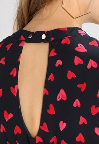 Tissu noir avec une petite ouverture à l'arrière, orné de motifs de cœurs rouges. Fermé par deux boutons métalliques au niveau du cou. Texture lisse.