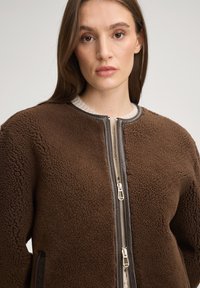 Brązowa kurtka polarowa z teksturowaną powierzchnią, zamkiem błyskawicznym oraz czarnymi akcentami wzdłuż zamka i kołnierza, wyposażona w boczne kieszenie.