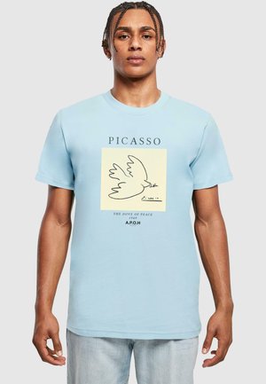 Šviesiai mėlyna marškinėliai su juoda baltojo balandžio piešiniu ant kreminės kvadratėlio, su užrašu "PICASSO" ir "TAIKOS BALANDIS 1949" apačioje.