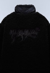 Veste en polaire noire avec un col montant et un logo embossé dans une teinte plus foncée. La texture est douce et pelucheuse.