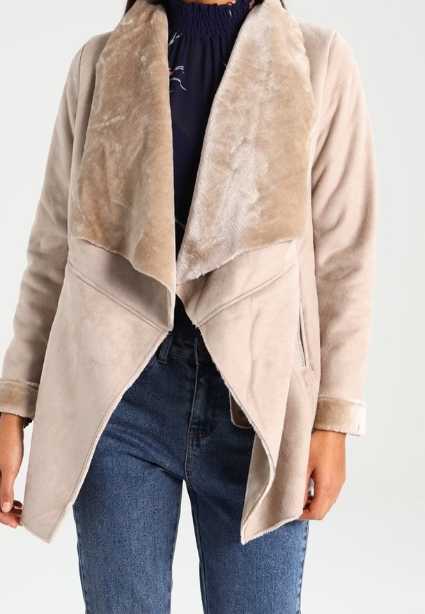 Beige jakke med blødt, struktureret stof og en kontrastfuld imiteret pelskrave. Har et asymmetrisk design og detaljer med lynlås i siden.